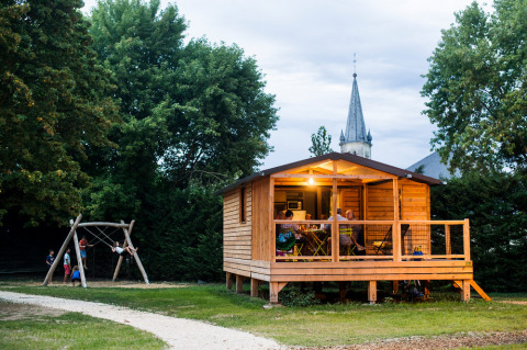Chalet Evasion hut bij Huttopia Les Châteaux, Frankrijk, met kinderen op de schommel en familie op het terras.