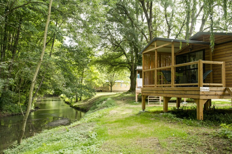 Cabaña de madera junto al río, Chalet Evasion Bord de Rivière, con terraza y rodeada de vegetación.