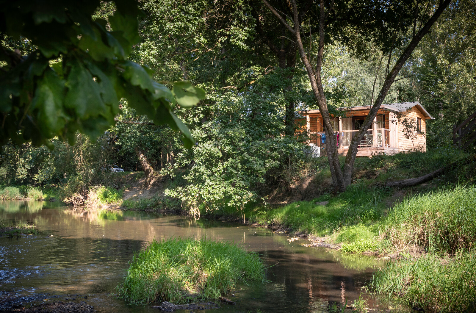 Houten chalet aan rivier, omgeven door groen bij Huttopia Les Châteaux in Frankrijk, rustieke natuur.