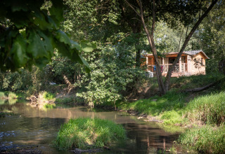 Cabaña de madera junto al río rodeada de vegetación en Huttopia Les Châteaux, Francia, escena tranquila.