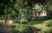 Cabaña de madera junto al río rodeada de vegetación en Huttopia Les Châteaux, Francia, escena tranquila.