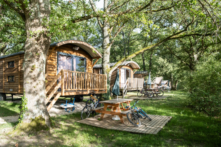 Roulotte cabins nestled in a wooded area with bikes, a picnic table, and lush green surroundings in summer.