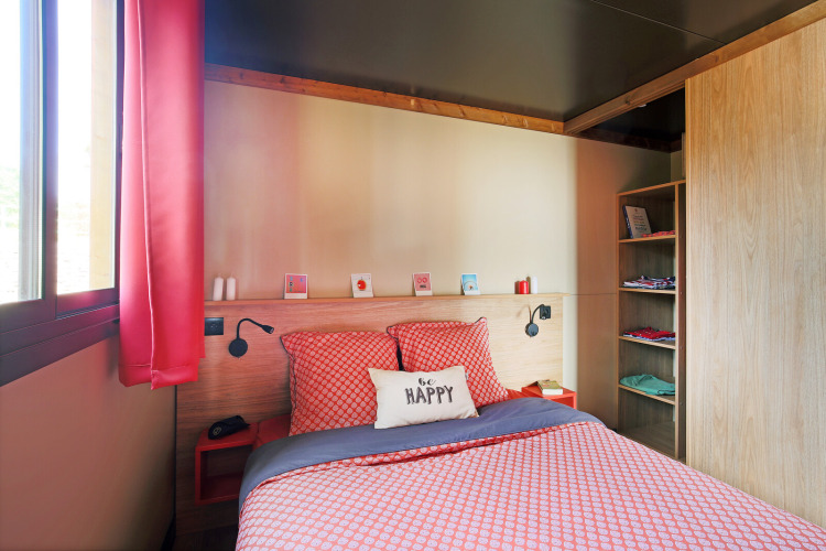 Cozy bedroom inside a Roulotte cabin at Huttopia Les Châteaux, France, featuring red decor and sunlight.