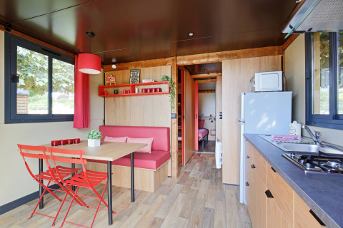 Hébergement glamping moderne avec kitchenette, coin repas, chaises rouges et grandes fenêtres lumineuses.