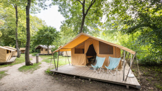 Classic safari tent with a wooden deck and two chairs, surrounded by trees in a forested campsite area.