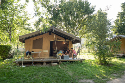 Tienda tipo safari Tent Sweet rodeada de árboles, con personas descansando en la terraza de madera.
