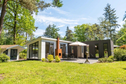 Moderne L-Pavilion lodge in een bosrijke omgeving, terras, eettafel, parasol en bijgebouw zichtbaar.