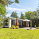 Moderne L-Pavilion lodge i en grøn skov, med terrasse, spisebord, parasol og ekstra bygning i baggrunden.