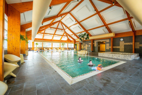 Piscina cubierta luminosa con vigas de madera, tumbonas y familias disfrutando en un alojamiento glamping.