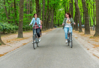 To kvinder cykler gennem en grøn skov på en sti ved et glamping-sted, en af dem har et barn bagpå.