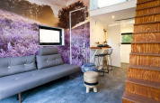 Modernes Tiny House Wohnzimmer mit Fototapete einer lila Wiese, grauem Sofa und Holztreppe in Holland.