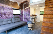 Sala moderna en mini casa con mural de campo de lavanda, sofá gris, escaleras de madera y cocina.