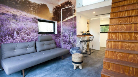 Moderne woonkamer in een tiny house met lavendelmural, grijze zetel, houten trap en kitchenette.
