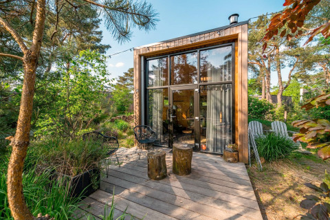 Moderne tiny house met grote ramen en houten terras midden in een weelderige, groene bosomgeving.