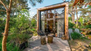 Tiny house moderne avec grandes vitrines et terrasse en bois dans un environnement forestier verdoyant.
