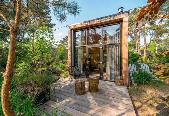 Moderne tiny house met grote ramen en houten terras midden in een weelderige, groene bosomgeving.
