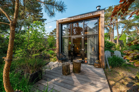 Modern tiny house with large windows and wooden deck surrounded by lush, green forest scenery.