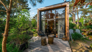 Moderne tiny house met grote ramen en houten terras midden in een weelderige, groene bosomgeving.