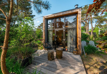 Moderne tiny house med store vinduer og træterrasse i frodig, grøn skovomgivelse om sommeren.