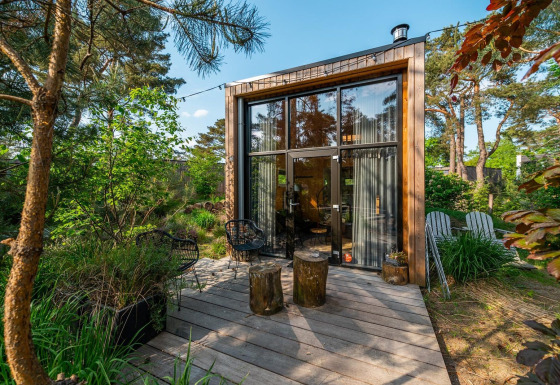Modern tiny house with large windows and wooden deck surrounded by lush, green forest scenery.