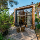 Moderne tiny house med store vinduer og træterrasse i frodig, grøn skovomgivelse om sommeren.