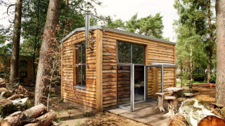 Petite maison nommée Tiny House en bois dans la forêt avec de grandes fenêtres et sièges extérieurs en rondins.