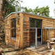 Et lille træhus ved navn Tiny House i skoven med store vinduer og udendørs siddepladser af træstammer.