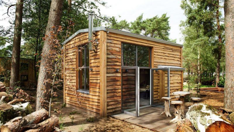 Una pequeña casa de madera llamada Tiny House en el bosque, con ventanas grandes y asientos de troncos afuera.