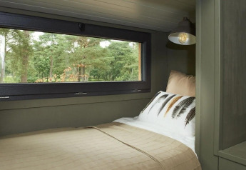 Knus bed bij groot raam in een tiny house op De Zanding, Nederland, met uitzicht op groene bomen.