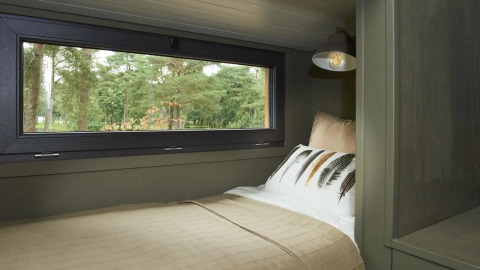 Cozy bed beside a large window in a tiny house at De Zanding, Netherlands, overlooking green trees.