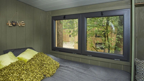 Acogedor dormitorio de tiny house con almohadas verdes, gran ventana y vista al bosque en De Zanding, Países Bajos.