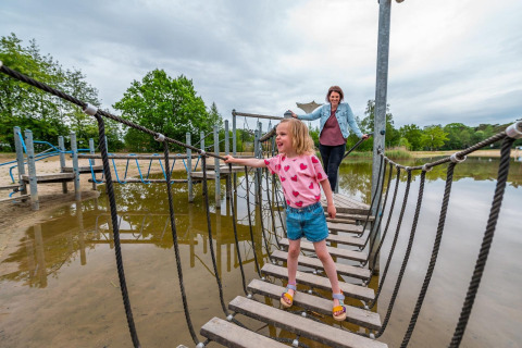 Niña cruza un puente de cuerda con un adulto detrás en un alojamiento glamping junto a un lago.