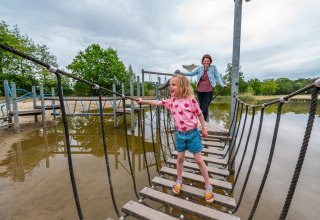 Niña cruza un puente de cuerda con un adulto detrás en un alojamiento glamping junto a un lago.