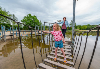 Niña cruza un puente de cuerda con un adulto detrás en un alojamiento glamping junto a un lago.