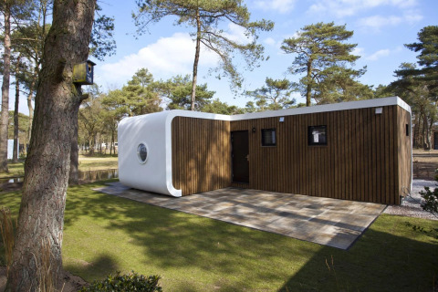 Modus L lodge with modern wooden façade and white rounded corner, set among pine trees in sunlight.
