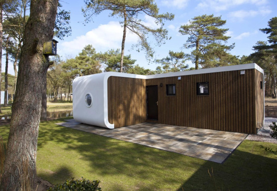 Modus L lodge with modern wooden façade and white rounded corner, set among pine trees in sunlight.