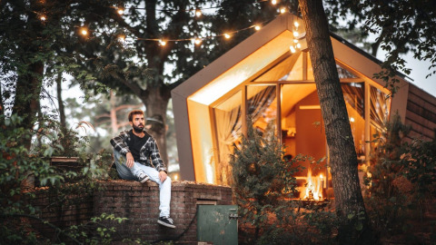 Mann sitzt draußen vor einer beleuchteten Lodge im Solo Retreat De Zanding in den Niederlanden am Abend.