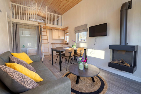 Moderne glampingindkvartering med hems, træloft, grå sofa, spisebord og vægmonteret pejs.