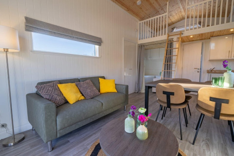 Alojamiento glamping moderno con sofá, mesa de comedor, altillo y decoración elegante en tonos naturales.