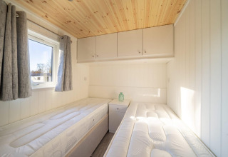 Lichte glamping slaapkamer met twee eenpersoonsbedden, raam, grijze gordijnen en houten plafond.