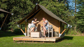 Dos personas brindando frente a una Glampingtent XS en Urban-Gardens Ieper, Bélgica, rodeados de naturaleza.
