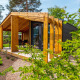 Panorama Cottage tiny house omgivet af træer og planter med en overdækket træterrasse i naturen.