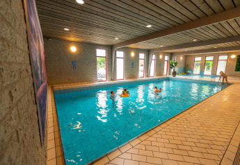 Piscina coperta in una struttura glamping, bambini che giocano in acqua, grandi finestre e piante decorative.