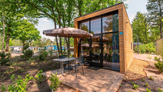 Moderne Tiny House lodge met houten gevel, hoge ramen en terras met tafel en parasol midden in het groen.