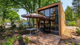 Tiny House lodge moderna con rivestimento in legno, grandi vetrate e terrazza con sedie e ombrellone.