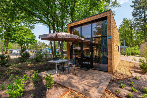 Moderne Tiny House lodge met grote glasramen, houten gevel en terras met parasol in een bosrijke omgeving.