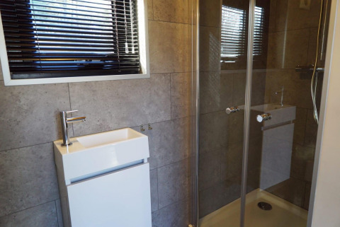 Baño moderno con ducha de vidrio, lavabo cuadrado pequeño y ventana con persianas horizontales negras.