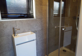 Baño moderno con ducha de vidrio, lavabo cuadrado pequeño y ventana con persianas horizontales negras.