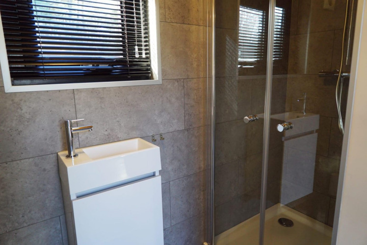 Baño moderno con ducha de vidrio, lavabo cuadrado pequeño y ventana con persianas horizontales negras.