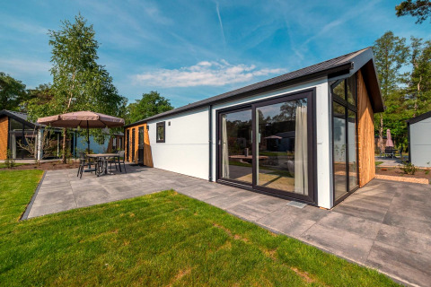 Lodge Velthorst moderne avec grandes baies vitrées, terrasse carrelée et mobilier de jardin, entouré d’arbres.